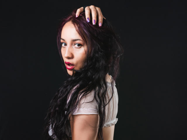 JessiicaDove con cabello negro largo y ondulado, lápiz labial rojo, esmalte de uñas morado, blusa blanca, mano en el cabello, mirando por encima del hombro, fondo negro..