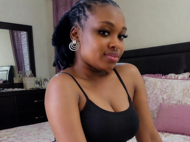 JuiceLipsx with dark skin, medium-sized boobs, black tank top, nose ring, spiral earrings, and curly black hair, smiling in a bedroom.