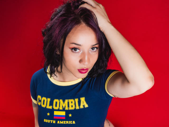 JessiicaDove with dark purple hair, blue "Colombia" shirt, yellow trim, hand in hair, red background, looking up with pink lipstick, slightly parted lips, and eyeliner.