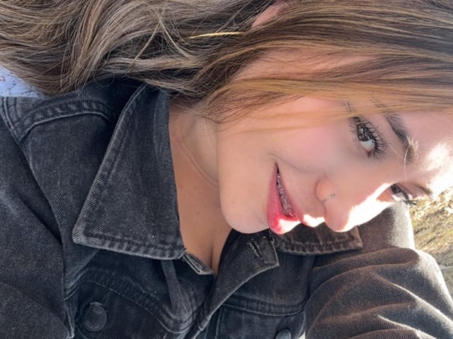 LorensDouce has light brown hair, green eyes, and red lipstick, smiling with her head tilted, wearing a dark denim jacket, showing her tits slightly, in a sunlit selfie.