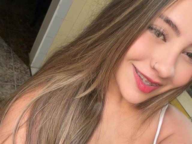 LorensDouce has long brown hair, a nose ring, and is smiling with pink lipstick and braces, wearing a white strap top. Her eyes are slightly closed.