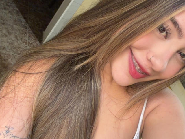 LorensDouce with long brown hair, smiling, pink lipstick, nose ring, white tank top, small tattoo on arm, visible breasts, soft light, slight tilt, relaxed, attractive, confident.