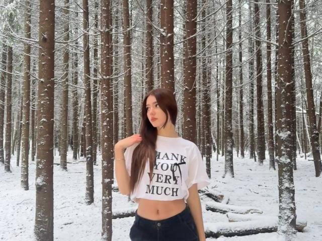 MaciePitcock se tient dans une forêt enneigée, vêtue d'un débardeur blanc et d'un pantalon noir. Elle a de longs cheveux bruns, une silhouette mince et un joli visage, et elle regarde sur le côté..