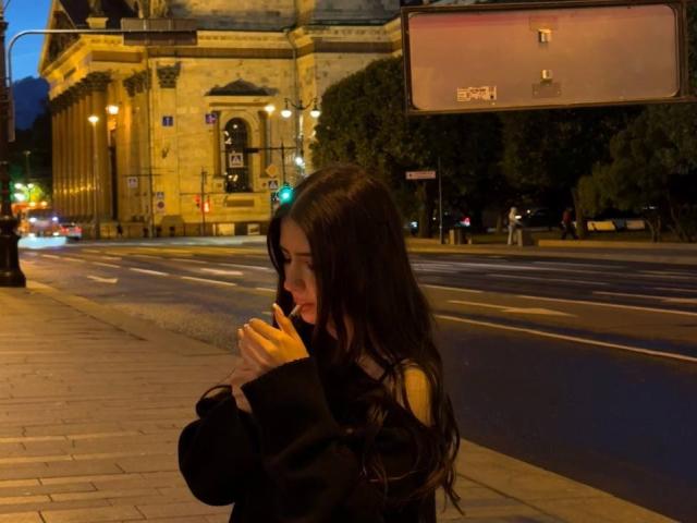 MaciePitcock with long dark hair, wearing a black top, holding a phone to her ear, standing on a sidewalk at night, with blurred car lights in the background.