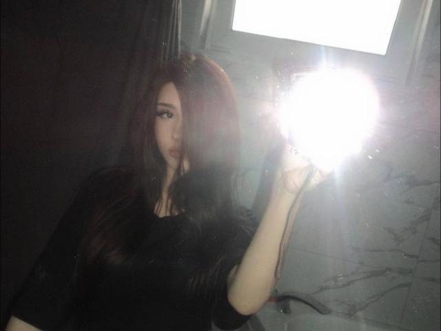 MaciePitcock with long dark hair, holding a bright flash in a mirror, wearing a black top, her tits slightly visible, in a dimly lit room.