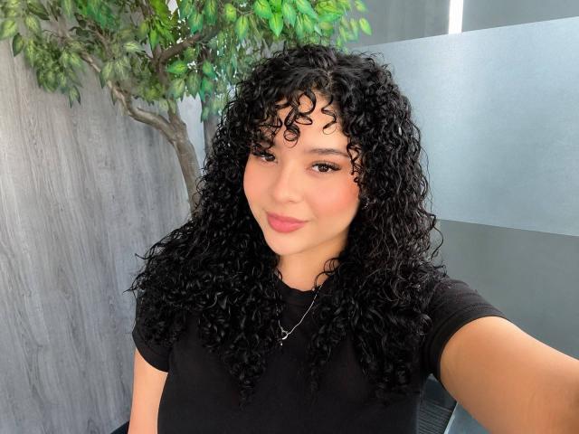 EmilyJay with curly black hair, wearing a black shirt, pink lips, and a small necklace, smiling slightly. Her tan skin and brown eyes are visible. She's taking a selfie.