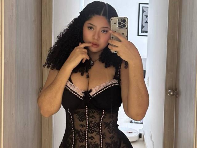 EmilyJay with curly black hair, wearing a black lace corset with pink lace trim, showing her tits, holding a phone, finger on her lip, mirror selfie.