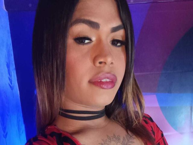 KeylaHotty69 with dark eyebrows, long straight brown hair, pink glossy lips, black choker, red patterned top, and tattoo on chest, looking slightly to the side, blue background.