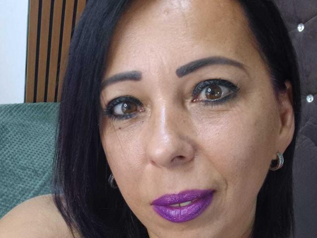 LauraSin tiene ojos marrón oscuro, cabello negro y lápiz labial morado. Lleva delineador de ojos negro, aros grandes y tiene una ligera sonrisa. Su piel es morena y se ve segura de sí misma..