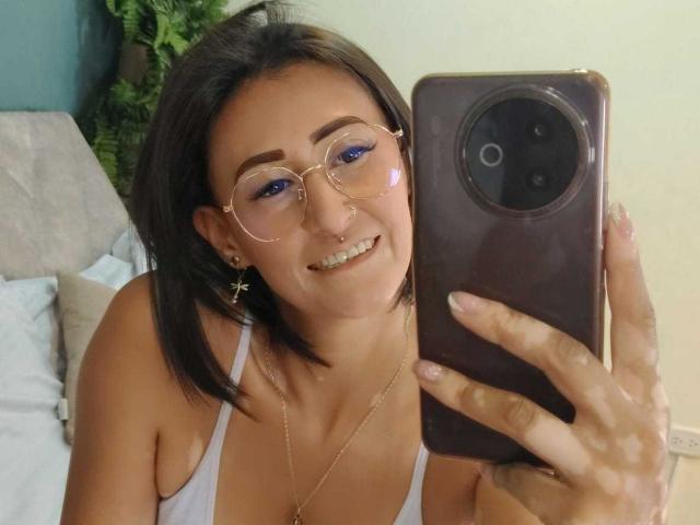 SharissWilliam with short brown hair, glasses, blue eyeshadow, cross earrings, nose ring, white tank top, smiling, holding a brown phone, visible breasts.