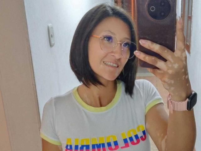 SharissWilliam with short black hair, glasses, and a smile, wearing a yellow-trimmed white tee, taking a mirror selfie, showing her tits and a pink wristwatch.