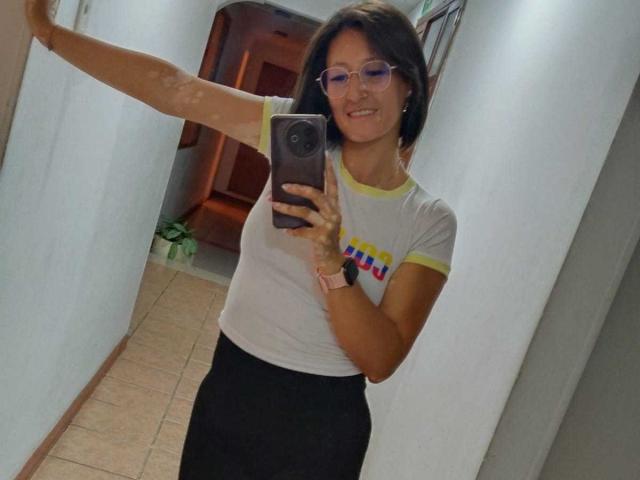 SharissWilliam with short brown hair, glasses, white t-shirt with yellow trim and colorful text, black skirt, taking a selfie in a hallway, smiling, right arm raised, holding phone.