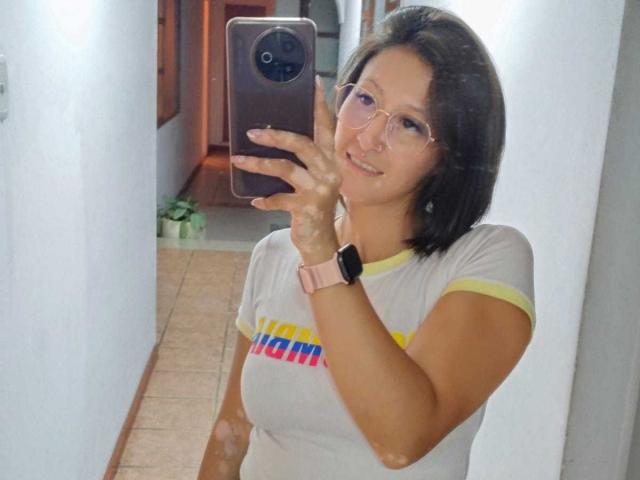 SharissWilliam taking a selfie, wearing glasses, pink watch, white shirt with yellow trim, holding a phone in a white hallway, smiling with short black hair.