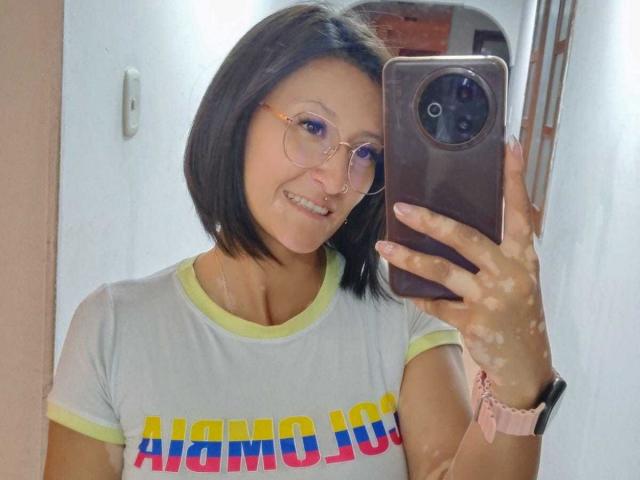 SharissWilliam with short black hair, glasses, smiling, wearing a yellow-green shirt, holding a phone, pink wristband, taking a mirror selfie, showing her boobs.