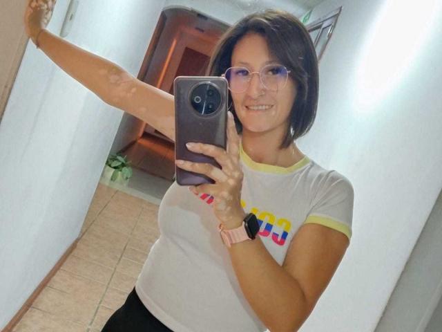 SharissWilliam with short brown hair, glasses, and pink wristwatch, taking a selfie with a smartphone, smiling, white shirt, yellow trim, holding phone with right arm raised.