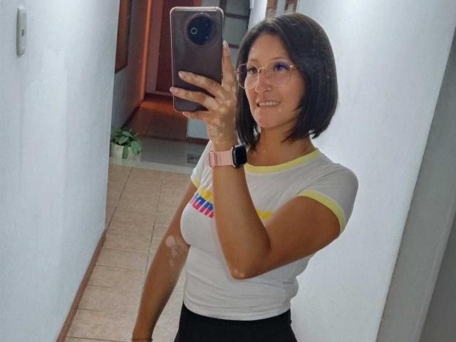 SharissWilliam with short black hair, glasses, white shirt with yellow trim, black pants, pink watch, holding phone, smiling, selfie in hallway, visible bores on arms.