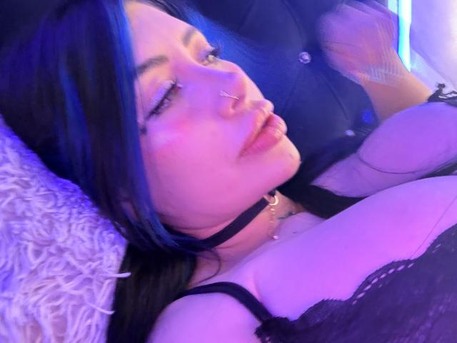 Kkole with blue highlights, nose ring, and pout, lying on a bed, wearing a black lace bra, exposing her big tits, and a black choker.