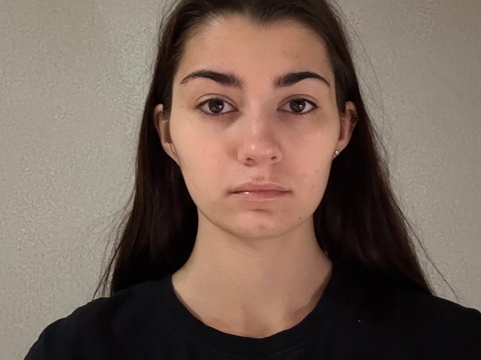 JudithNorick has long brown hair, dark eyebrows, brown eyes, and is wearing a black shirt with a neutral expression. Her skin is light and she has small earrings.