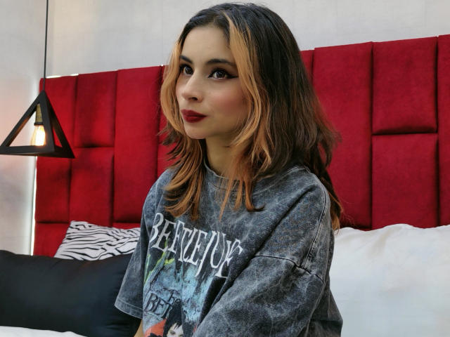 Amuneet with long, wavy brown hair and red lipstick, wearing a gray, graphic-print, oversized shirt, sitting on a bed with a red, tufted headboard.