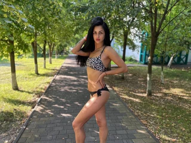 EvaClarkX in a leopard print bra and black string panties, standing on a brick path, hand in hair, looking at camera, green trees, sunny, cute, sexy, small butt.