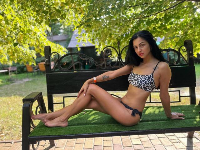 EvaClarkX with dark hair, wearing a leopard print bikini, sits on a green bench with her legs crossed, showing her ass and small tits, in a sunny park.
