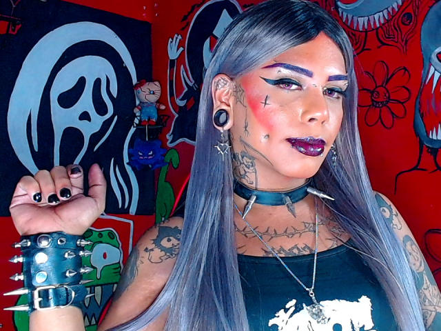 MarcelineDarketa con capelli grigi, rossetto scuro, unghie nere, tatuaggi, una collana nera con borchie, un braccialetto con borchie e orecchini, in una foto in stile punk. Sfondo rosso con arte surreale..
