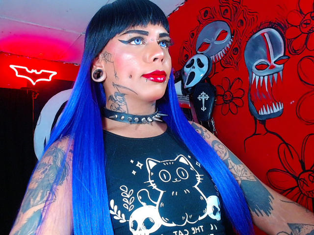 MarcelineDarketa con capelli blu, tatuaggi, rossetto rosso, maglietta con un gatto nero, collana con borchie, piercing alle orecchie, che guarda verso l'alto, sfondo rosso e nero con facce di teschio. Il seno è visibile, i seni sono leggermente coperti..