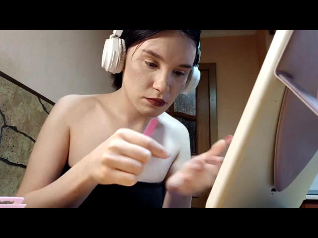 CherWinger with pink dildo in hand, wearing white headphones, focused on computer screen, topless with bare shoulders, brown hair, light skin, serious expression, indoors.