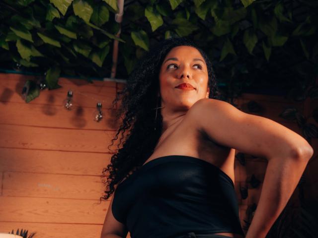 Samannttha, con cabello negro rizado, lápiz labial rojo y un top negro sin tirantes, mirando hacia arriba con una ligera sonrisa, mostrando sus senos y una postura segura. Fondo de madera cálida..
