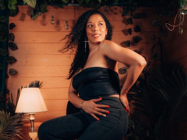 Samannttha with curly black hair, wearing a black strapless top and dark jeans, smiling, hand on hip, red nails, standing by a lamp, wood wall, and leafy plants.
