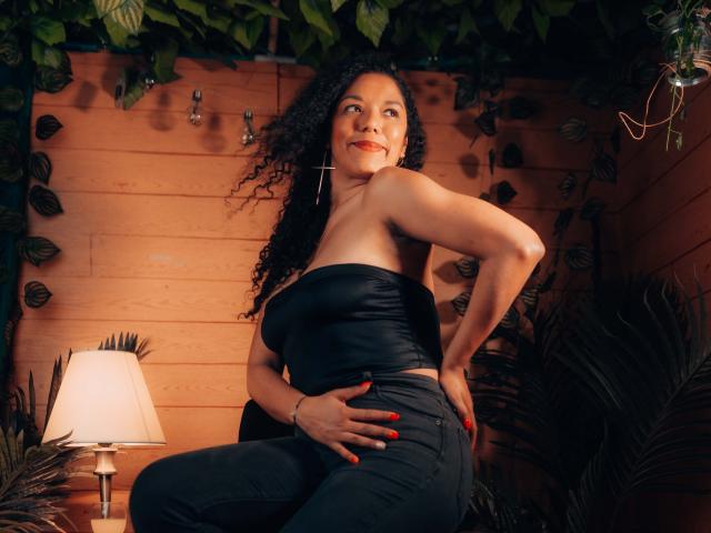 Samannttha, con cabello negro rizado, vestida con una blusa negra sin tirantes, jeans negros y aretes colgantes, sonriendo, con las manos en las nalgas, de pie en una habitación con luz cálida y una lámpara..