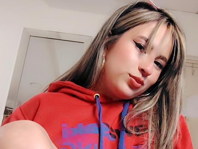 AmiiRosse with pouty lips, wearing a red hoodie, blue drawstrings, and a pink headband, has blonde highlights in her long hair, and is looking slightly to the side.