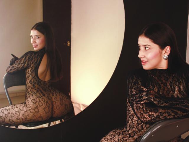 EmmaLombardi in a sheer, black lace bodysuit with her back to a mirror, showing her ass. She's looking over her shoulder, smiling, with dark hair and earrings.