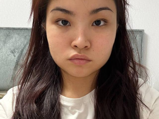 LeoraHardeman's face with medium brown skin, dark brown straight hair, small nose, and serious expression, wearing a white shirt, looking straight at the camera.