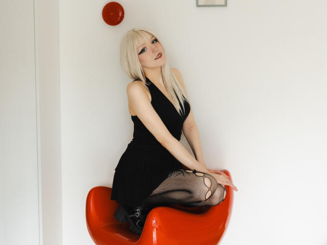NikaDreamOne with blonde hair, black dress, and sheer black pantyhose, sitting on an orange chair, showing her tits and ass, wearing black boots, in a white room.