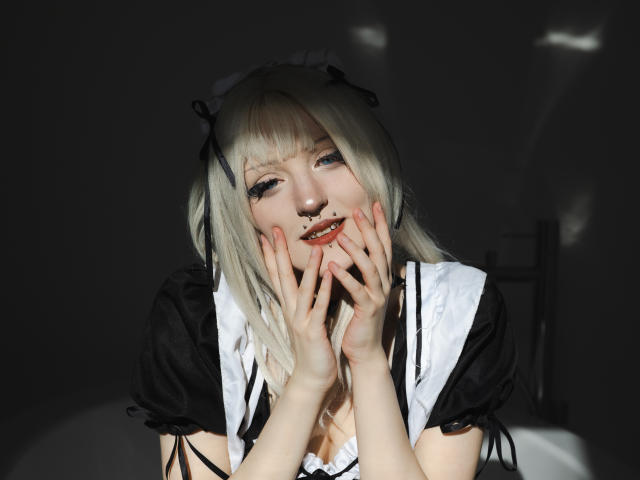 NikaDreamOne with pale skin, white blonde hair, black and white maid outfit, hands near face, smiling, red lipstick, black eye makeup, fingers with pink nail polish, dark background.