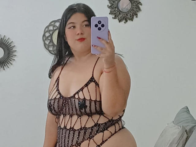 SultanaLm, avec des cheveux noirs, du rouge à lèvres et une peau de couleur moyenne, portant une lingerie noire transparente en maille laissant entrevoir ses seins, tenant un téléphone violet, prenant un selfie devant un miroir..