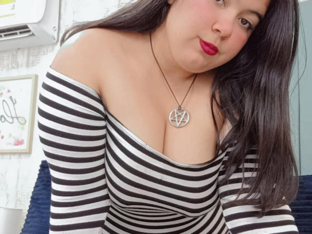 SultanaLm with large tits, wearing a black and white striped off-shoulder top, red lipstick, and a pentagram necklace, leaning forward, long dark hair, slight smile.