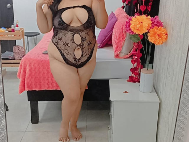 SultanaLm en lingerie noire en dentelle avec découpes, montrant ses grandes seins et ses fesses rebondies, debout dans une chambre avec un lit rose et des fleurs orange sur une table de chevet..