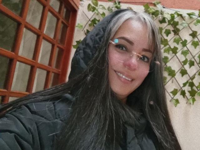 LailaCastillo with long black hair, smiling, wearing a black puffy jacket, glasses, and a hood, standing outside near a red wooden window. Her tits are not visible.