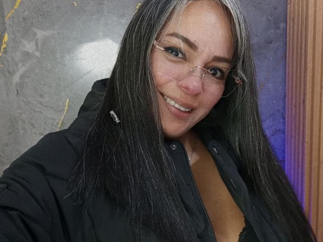 LailaCastillo has long black hair, glasses, and a smiling face, wearing a black jacket with a visible black bra underneath, showing her boobs. She's posing for a selfie.