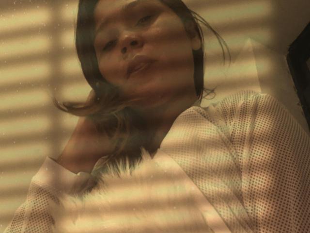 SophiaQueens tiene una cara linda con cabello castaño, vestida con una camiseta blanca de malla, mirando ligeramente hacia abajo, con franjas de luz y sombra sobre ella. Sus labios están ligeramente entreabiertos..