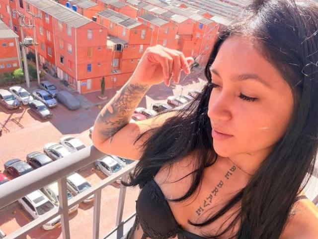 MiaaVixen with black hair, tattoos on arm and chest, black bra, standing on balcony, looking down, sunlight, cars in parking lot, orange apartment buildings in background.