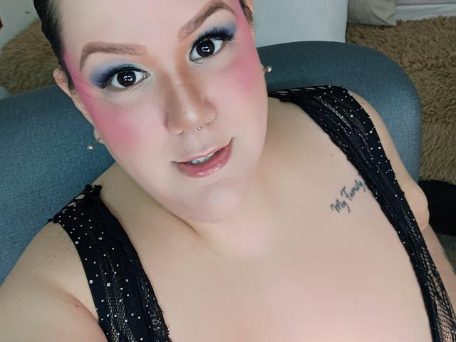 GiiannaMorrys with pink blush, blue eyeshadow, nose piercing, black sparkly dress, visible tattoo, and small boobs, looking up with a slight smile.