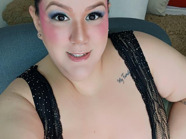 GiiannaMorrys with pink blush, blue eye makeup, black lace top, nose ring, small earrings, tattoo on right shoulder, smiling, fair skin, brown hair, and chubby, sitting on grey chair.
