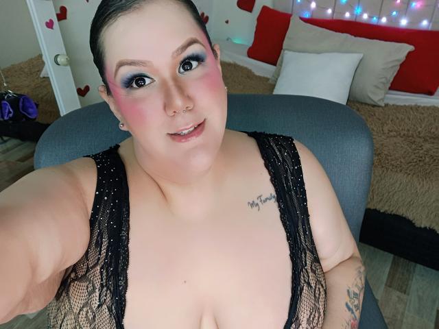 GiiannaMorrys has pale skin, black lace top, pink blush, blue eyeshadow, nose ring, tattoo on right chest, and a smile, showing her tits in a selfie.