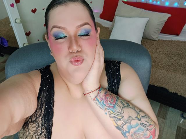 GiiannaMorrys, plus-sized with blue eyeshadow, pink blush, nose ring, black lace top, hand on cheek, tattoo on chest, puffy lips, sitting on gray chair, bedroom.