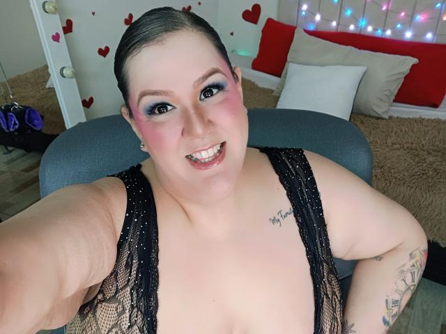 GiiannaMorrys, plus-sized with pink eyeshadow and black lace top, showing off her tits and tattoo, smiling with arms on gray chair, dark hair pulled back.