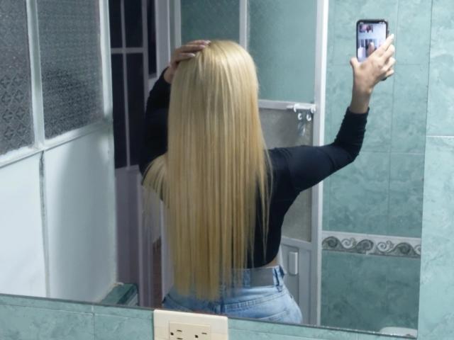TanniaMoon, con lunghi capelli biondi, una maglietta nera e jeans blu, sta scattando un selfie allo specchio, mostrando la sua schiena, con una mano sulla testa e il telefono nella mano destra..