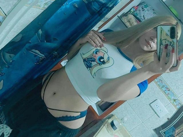 TanniaMoon, con i capelli biondi, indossa una maglietta bianca a raglan, un laccio nero in pizzo e calze blu che arrivano all'altezza coscia, mostrando il suo ventre piatto e i suoi piccoli seni in un selfie allo specchio..